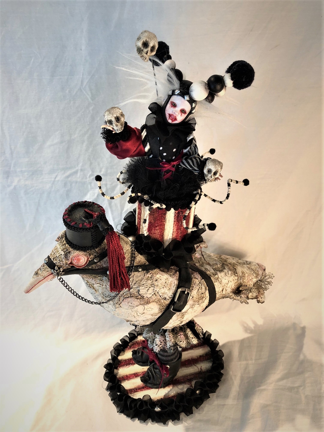 mixed media miniature gothic costumed circus performer juggles skulls and rides a lace-covered albino crow toy wearing striped stockings, boots and a fez standing on a painted wooden platform