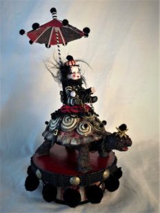 mixed media striped miniature gothic circus performer in black white and red stripes, shaded by a parasol rides a hand-painted vintage toy turtle on a painted wooden platform