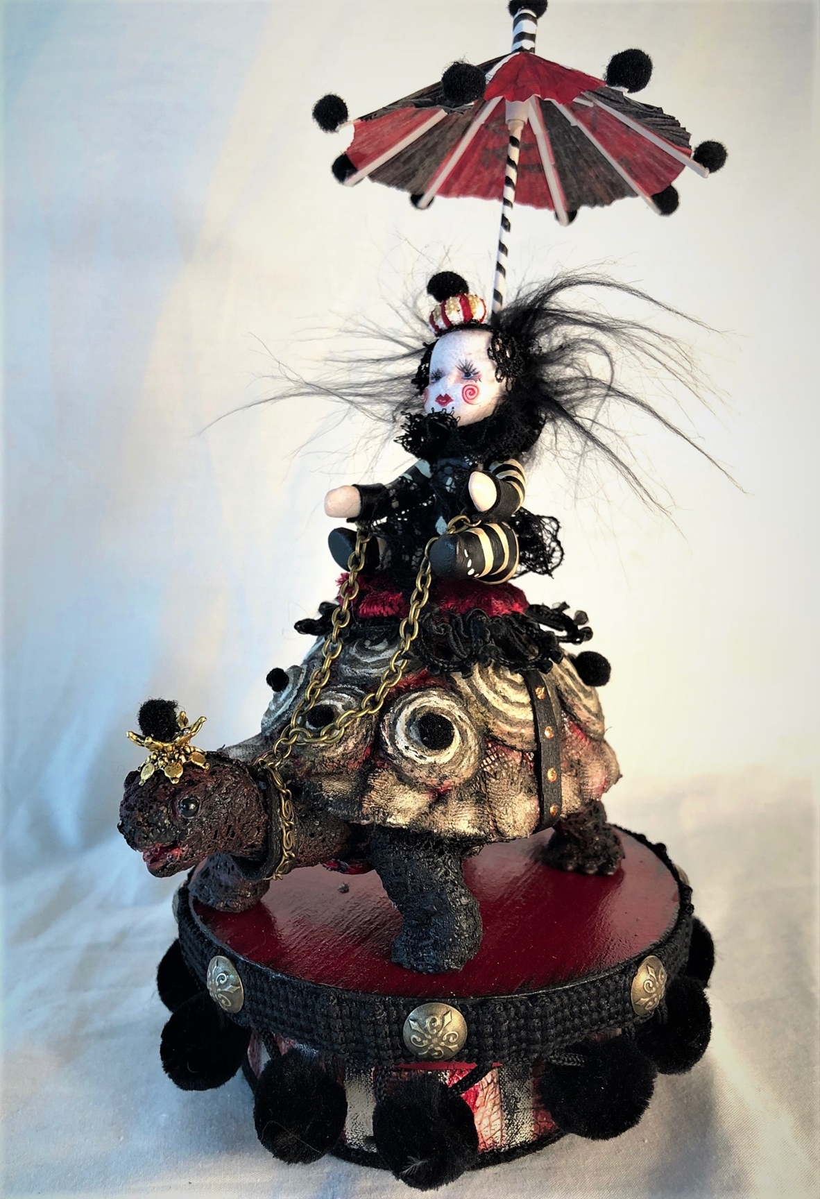 mixed media striped miniature gothic circus performer in black white and red stripes, shaded by a parasol rides a hand-painted vintage toy turtle on a painted wooden platform