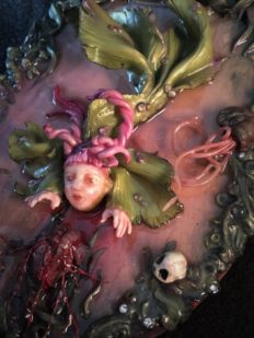 resin encased mixed media assemblage hand painted doll-faced green and pink fish creature reaching for a heart at water's edge.