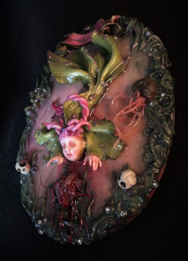 resin encased mixed media assemblage hand painted doll-faced green and pink fish creature reaching for a heart at water's edge.