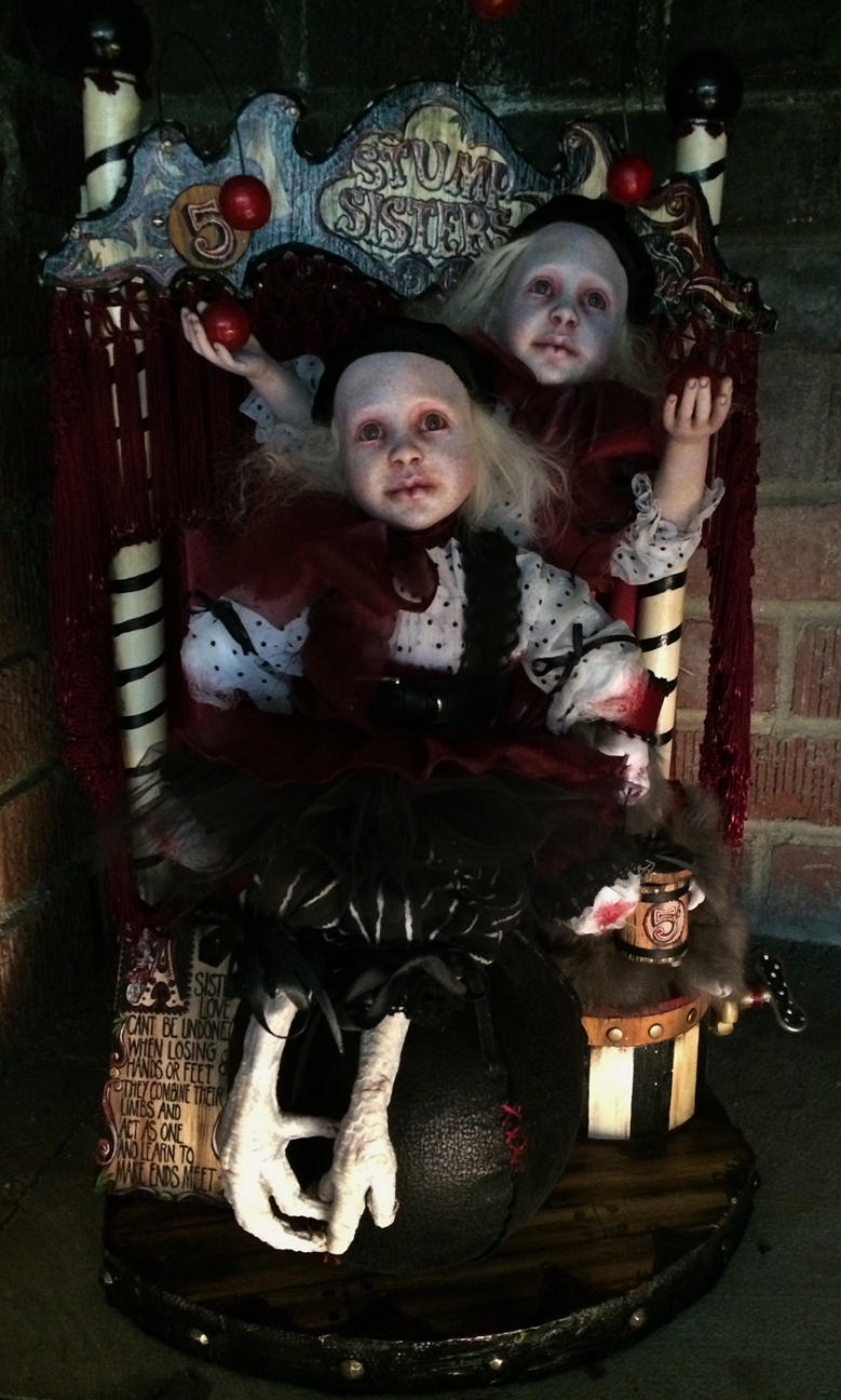 gothic artdolls of a balancing sister act one doll has no hands or feet and balances on her sister's lap with taxidermy bird feet on a wooden platform with hand-painted signs