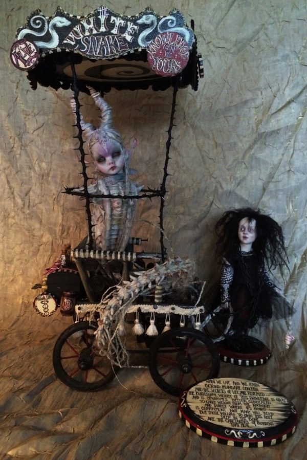 snake-woman hybrid doll freakshow sideshow in a circus cart with hand-painted lettered circus signs and a small gothic doll with black hair in a black dress standing to the side