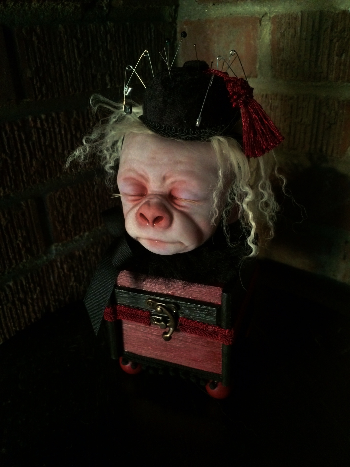 pale, albino gorilla face doll head with closed eyes and blond hair wearing a black pin cushion with a red tassel on his head set on top of a burgundy and black wooden box