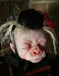 close-up pale, albino gorilla face doll head with closed eyes and blond hair wearing a black pin cushion with a red tassel on his head