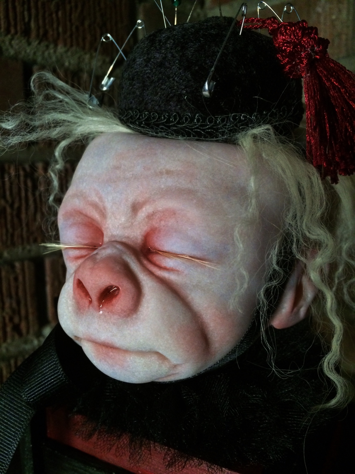 close-up pale, albino gorilla face doll head with closed eyes and blond hair wearing a black pin cushion with a red tassel on his head