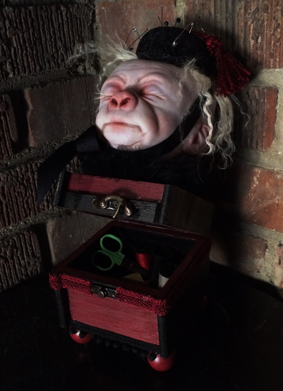 pale, albino gorilla face doll head with closed eyes and blond hair wearing a black pin cushion with a red tassel on his head set on top of an open burgundy and black wooden box with sewing notions inside the box