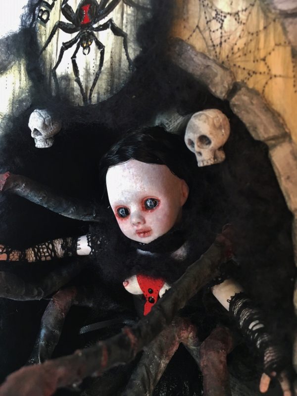 close-up shadowbox mixed media assemblage of a gothic black widow themed doll with 8 legs pale with black hair and red hour glass she is inside a carved box with hand-painted wooden signs