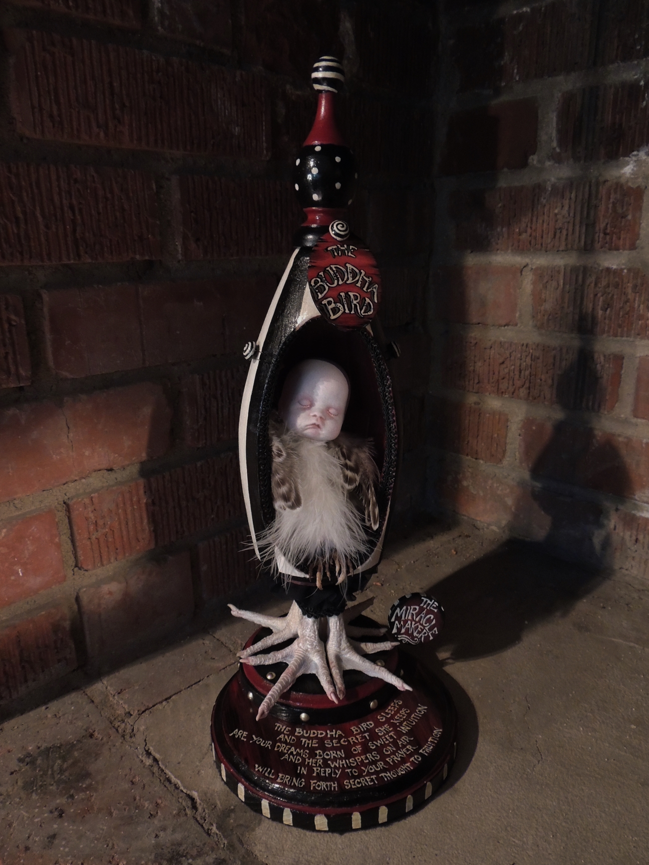 sleeping baby doll with feathered bird body in an egg shaped containter with taxidermy bird feet standing on red and black hand-painted wooden platform with lettered verse