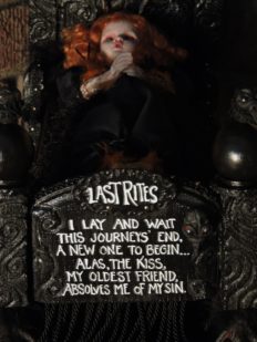 close-up of a miniature red-headed doll on her dark deathbed with a hand painted poem on the footboard