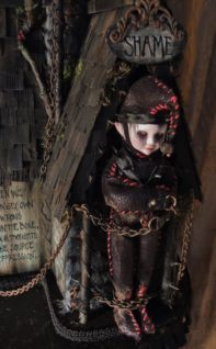 close-up of a miniature goth elf chained in front of a door standing under a sign that says Shame