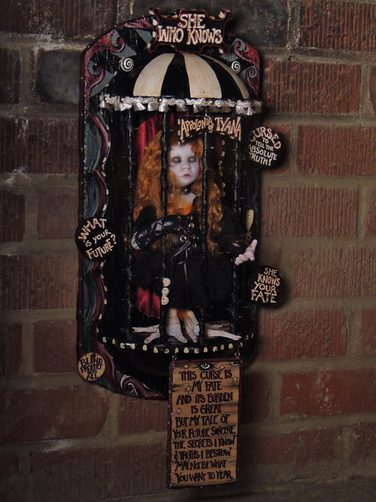 gothic redheaded blind seer artdoll with taxidermy birdfeet trapped in a circus themed black thorny cage with hand-painted signs on it wall-mounted