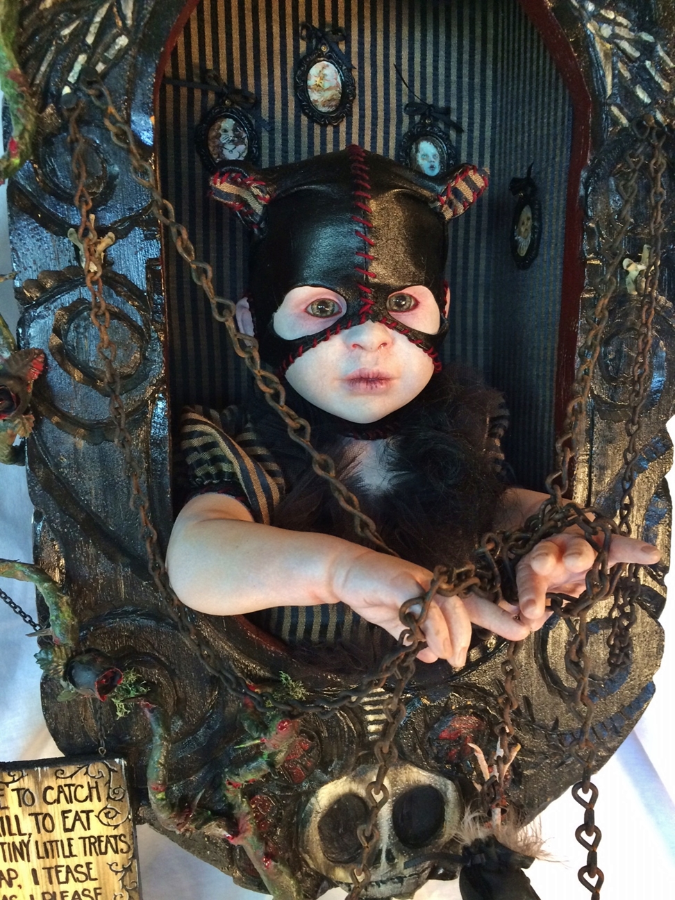 close-up of gothic artdoll wearing black leather cat mask holding chains inside carved shadowbox with skull