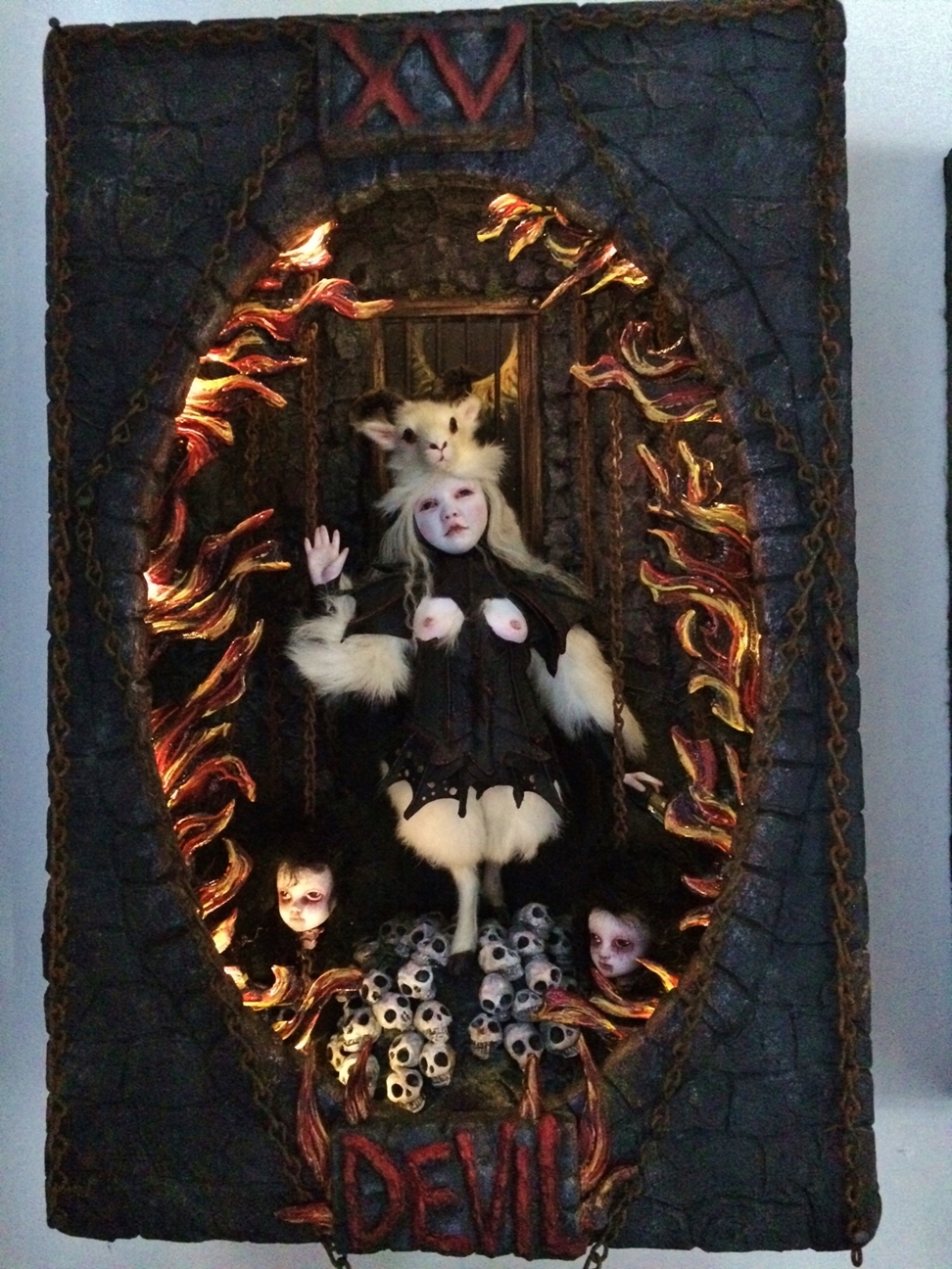 miniature shadowbox diorama with hellscape scene inside a tiny devil doll with a goat headdress and black leather vest surrounded by tiny skulls doll heads and hand painted flames