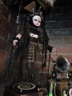 gothic steam punk art doll stands on a circular platform behind a steampunk machine contraption with phonograph