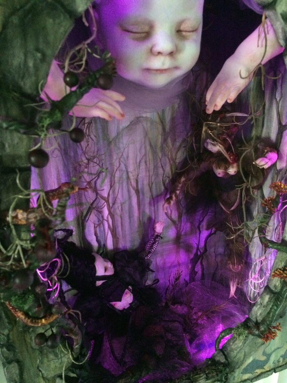 miniature shadowbox diorama lit with purple led light with two tiny gothic dolls reaching for each other in a forest setting with a bigger doll above them