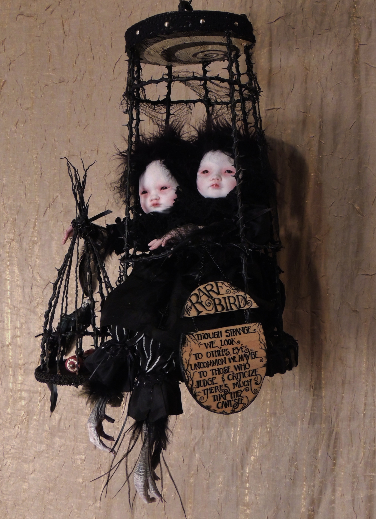 taxidermy mixed media art doll assemblage art piece featuring black feathered pale asian doll-faced conjoined twins sitting in a hanging cage.