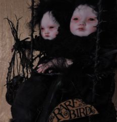 close-up taxidermy mixed media art doll assemblage art piece featuring black feathered pale asian doll-faced conjoined twins sitting in a hanging cage.