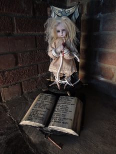 a blond artdoll with taxidermy bird feet is bound to a carved wooden door above a book with verse telling her story music box