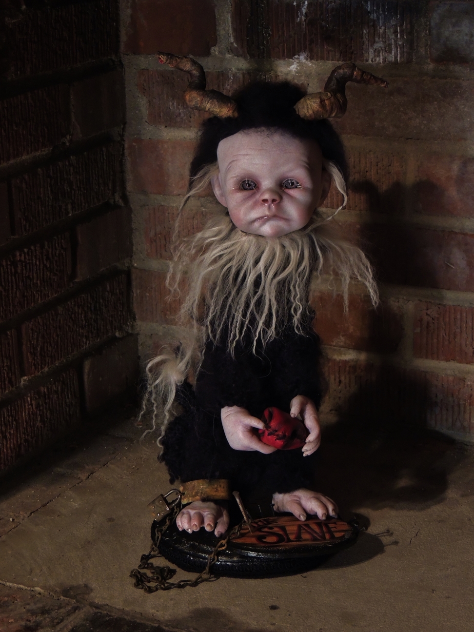 mixed media artdoll has hand-painted vinyl dollhead scrunched face with hand-sculpted horns, covered in black mohair fur, holds his heart in his hands